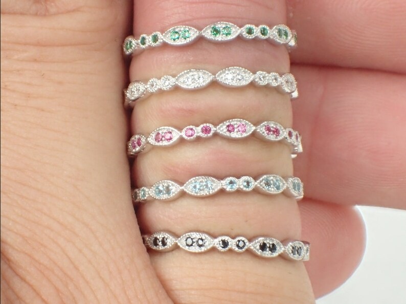 May include: Five delicate silver rings with different colored gemstones. Each ring features a row of small, oval-shaped gemstones set in a silver band. The gemstones are green, white, pink, blue, and black.