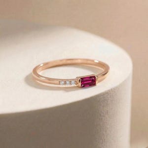 May include: Rose gold ring featuring a rectangular, deep red gemstone and three small, clear accent stones. The ring has a delicate band and a minimalist design, perfect for everyday wear or special occasions.