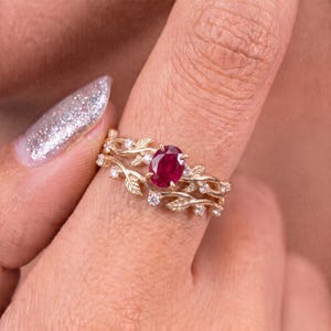 Könnte beinhalten: Ein Goldring mit einem zentralen, oval geschliffenen, tiefroten Rubin. Die Ringschiene ist mit einem strukturierten Weinreben- und Blattmotiv gestaltet, das mit kleinen, runden weißen Steinen entlang der Zweige besetzt ist und eine botanische Optik erzeugt.