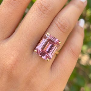 Può includere: Un anello d'oro con una grande gemma rosa rettangolare. La gemma, con taglio a smeraldo, è tenuta in posizione da quattro griffe d'oro. L'anello è su un dito, con uno sfondo verde sfocato.