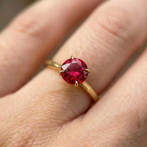 May include: A gold ring featuring a large, round, vibrant red gemstone. The gemstone is held in place by four prongs, and the band is a simple, polished gold. The ring is shown on a finger.