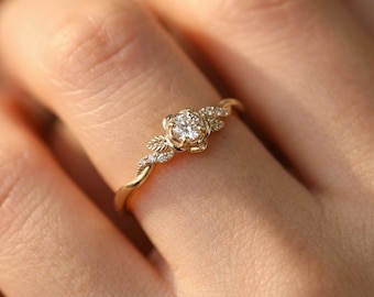 Könnte beinhalten: Ein goldener Ring mit floralem Design, zentralem Diamanten und kleineren Akzentsteinen. Das Band hat ein gedrehtes Design mit blattartigen Details. Der Ring befindet sich an einem Finger und unterstreicht seine filigrane Handwerkskunst und seinen eleganten Stil.