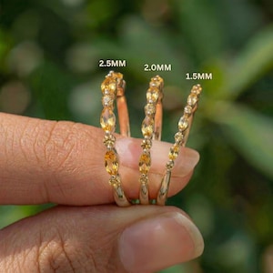 May include: Three gold rings with yellow gemstones are displayed. The rings are labeled 2.5MM, 2.0MM, and 1.5MM. Each ring features a series of oval and round yellow stones set in a gold band. The rings are held against a blurred green background.