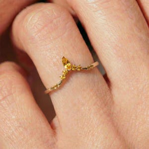 Può includere: Un anello color oro con un design curvo, caratterizzato da una gemma gialla centrale a forma di marquise e pietre gialle rotonde più piccole. L'anello è indossato su un dito, mostrando il suo stile delicato ed elegante.