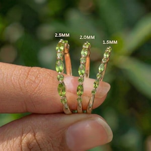 May include: Three gold rings with green gemstones. The rings are labeled 2.5MM, 2.0MM, and 1.5MM. The gemstones are oval and round, set in a row along the band. The background is a blurred green.