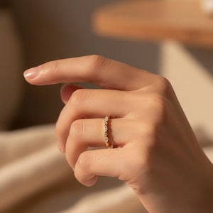以下が含まれることがあります： 小さな透明な宝石が並んだゴールドリング。指にはめられ、背景はぼやけています。リングのデザインは繊細でエレガントで、普段使いにも特別な機会にも適しています。