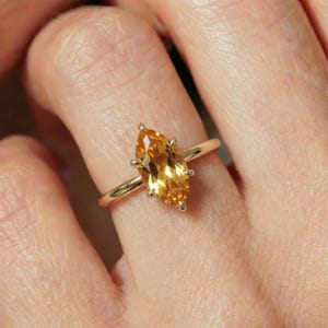 May include: A gold ring featuring a marquise-cut, golden-yellow gemstone. The gemstone is set with prongs, and the band is a simple, polished gold. The ring is shown on a finger.