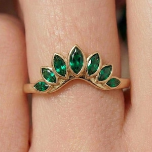 Może przedstawiać: Złoty pierścionek z serią markizowych, żywych zielonych kamieni szlachetnych, ułożonych w zakrzywiony, przypominający koronę wzór. Oprawa pierścionka jest z polerowanego złota, a kamienie szlachetne są blisko siebie osadzone.