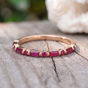 May include: A rose gold band ring featuring an alternating pattern of baguette-cut and round-cut red rubies set along the top half of the metal band. The ring has a polished finish and a slim profile, showcasing the vibrant red gemstones clearly.