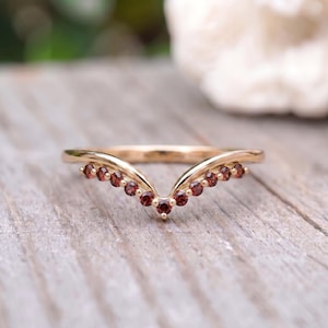 Pode incluir: Um anel em forma de V feito de metal dourado polido, apresentando uma série de pequenas pedras preciosas granadas redondas e vermelho-escuras cravadas ao longo da banda estilo chevron. O design segue um arco simétrico com as pedras incrustadas.