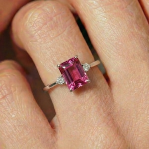 May include: A silver ring featuring a large, rectangular, vibrant pink gemstone as the centerpiece. Two smaller, round, clear gemstones flank the main stone. The ring is set on a finger, showcasing its elegant design.