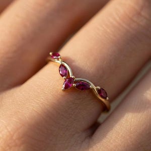 May include: A gold ring with a curved design, set with marquise-cut ruby gemstones. The ring's band is thin, and the rubies are a vibrant red color. The ring is shown on a finger.