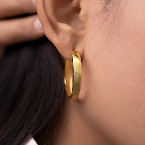 Puede incluir: Un par de pendientes de aro de oro. Los pendientes son gruesos y tienen una forma ligeramente redondeada.