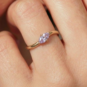 May include: A gold ring featuring a single marquise-cut light purple gemstone set in a prong setting. The band has a smooth, polished finish and a slightly curved design that tapers toward the center stone, creating a delicate and refined appearance.