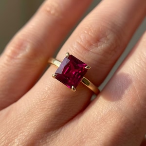 May include: A gold ring featuring a large, square-cut, deep red gemstone. The gemstone is held in place by four gold prongs. The ring's band is a simple, polished gold. The ring is a piece of jewelry.