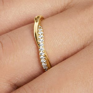 Könnte beinhalten: Ein goldfarbener Ring mit einem gedrehten Design. Eine Seite des Rings ist mit einer Reihe kleiner, klarer Edelsteine verziert. Der Ring ist vor einem verschwommenen Hintergrund platziert.