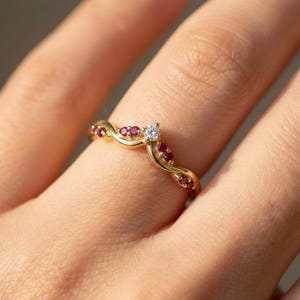 May include: A gold ring with a wave design, set with small red gemstones and a single, central clear gemstone. The ring is shown on a finger, highlighting the intricate details of the jewelry.