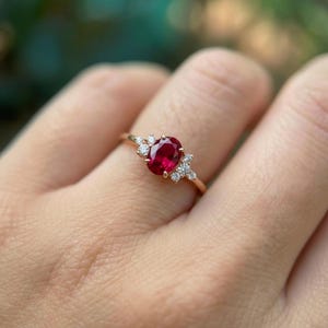 May include: A gold ring featuring a large oval-cut ruby gemstone as the centerpiece, flanked by clusters of smaller, round diamonds. The ring's band is a delicate gold color, and the gemstones are set in a prong setting.