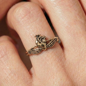 Op de afbeelding: Een goudkleurige metalen ring met een centraal anatomisch hartontwerp geflankeerd door twee skeletachtige handen die om de band heen grijpen. De ring heeft een gepolijste afwerking met gedetailleerde texturen op het hart en de vingerkootjes.