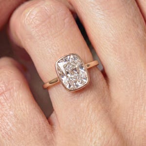 May include: A rose gold ring featuring a large, rectangular cushion-cut center stone with rounded corners. The gemstone is set in a smooth, polished bezel setting that encircles the entire perimeter of the stone, connected to a thin, plain band.