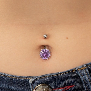 May include: A belly button ring with a silver barbell and a purple flower-shaped charm. The charm has a large, round center stone surrounded by smaller stones. The ring is shown against a background of skin and denim jeans.