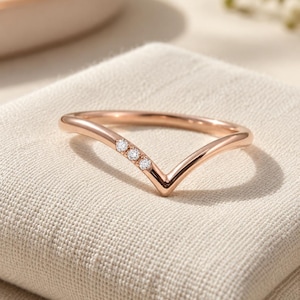 May include: A rose gold V-shaped ring featuring three small, clear, round-cut stones set along one side of the chevron band. The metal has a polished, reflective finish, and the design creates a pointed silhouette that sits centered on the band.