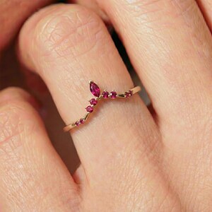 May include: A gold-colored ring with a curved design, set with small, vibrant red gemstones. The ring features a marquise-cut ruby at the top, with smaller round rubies along the curve. A delicate and elegant piece of jewelry.