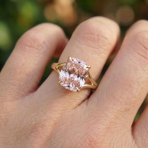 Puede incluir: Un anillo de oro con una gran gema de corte cojín de color rosa claro. La banda del anillo tiene un diseño dividido, y la gema se sujeta con cuatro garras. El anillo se muestra en un dedo.
