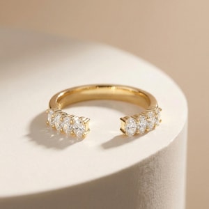 May include: A gold-colored ring with an open design, featuring a row of five clear, oval-cut gemstones on each side. The ring is set on a white surface, with a soft, neutral background.