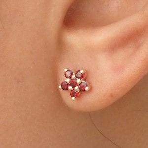 Può includere: Un paio di orecchini a perno in oro con un design a forma di fiore rosso. Il fiore ha otto petali ed è incastonato con piccole pietre preziose rosse.