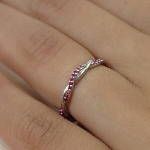 May include: A silver-colored ring with a twisted design, adorned with small, vibrant pink gemstones. The ring is shown on a person's finger, highlighting its delicate and elegant appearance. The gemstones are evenly spaced along the ring's surface.