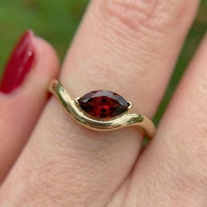 Puede incluir: Un anillo de oro con una gema roja oscura con corte marquesa. La banda del anillo se curva con gracia, sosteniendo la piedra. La gema es el punto focal, mostrando su rico color y diseño facetado. El anillo es una pieza de joyería.