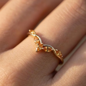 Może przedstawiać: Złoty pierścionek o zakrzywionym kształcie, z kilkoma pomarańczowymi kamieniami szlachetnymi o szlifie markizy. Pierścionek jest umieszczony na rozmytym tle, podkreślając szczegóły biżuterii.