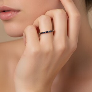 May include: A gold band ring featuring a series of rectangular, dark blue gemstones. The ring is set with small, round gold accents between the blue stones. The ring is shown on a person's finger.