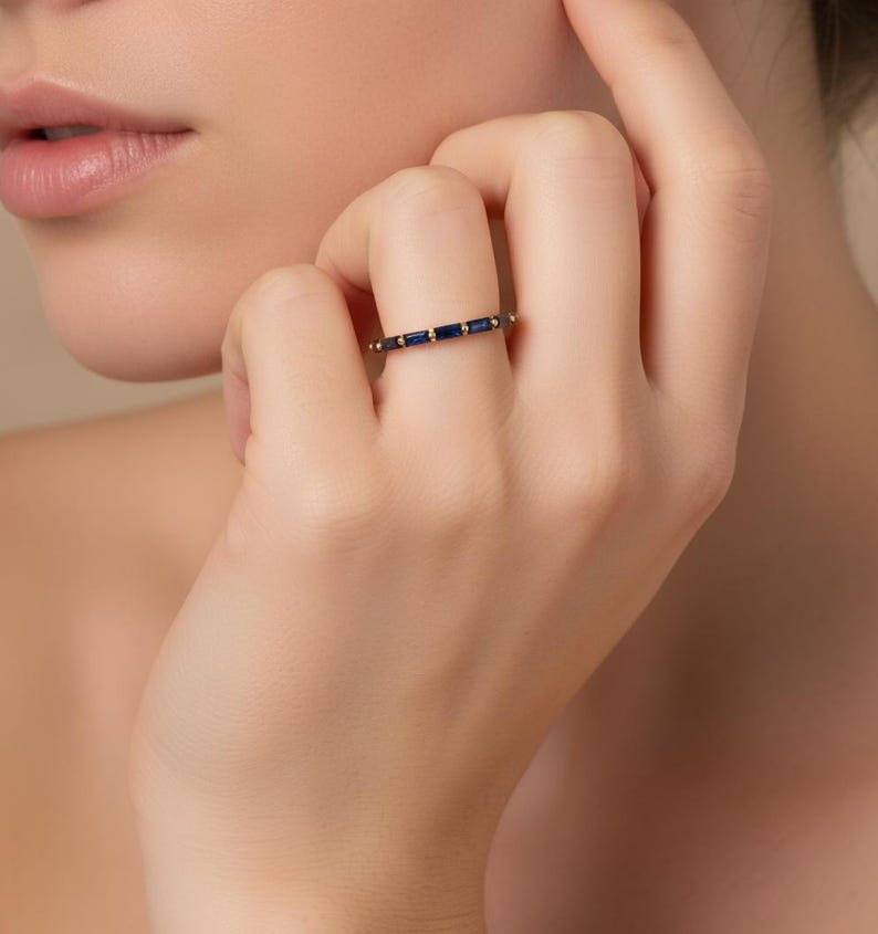 May include: A gold band ring featuring a series of rectangular, dark blue gemstones. The ring is set with small, round gold accents between the blue stones. The ring is shown on a person's finger.