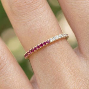 May include: A gold band ring set with alternating red rubies and white diamonds. The ring is on a finger, with the gemstones encircling the band. The ring's design is delicate and elegant, suitable for everyday wear or special occasions.