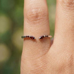 Könnte beinhalten: Ein silberfarbener offener Ring mit drei kleinen, ovalen, dunkelroten Edelsteinen auf jeder Seite. Der Ring ist vor einem verschwommenen grünen Hintergrund platziert.