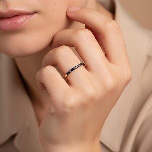 May include: A gold ring with a series of dark blue rectangular and round gemstones. The ring is worn on a finger, showcasing the jewelry's design and color contrast. The ring's band is a thin, polished gold.