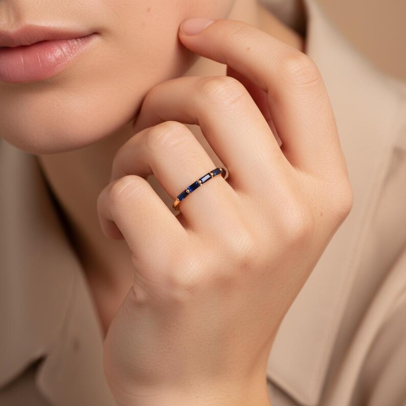 May include: A gold ring with a series of dark blue rectangular and round gemstones. The ring is worn on a finger, showcasing the jewelry's design and color contrast. The ring's band is a thin, polished gold.