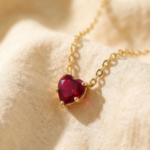 May include: A gold-colored necklace with a heart-shaped, deep red gemstone pendant. The pendant is set in a gold bezel and hangs from a delicate chain. The necklace is displayed on a cream-colored fabric.