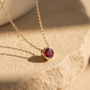 Pode incluir: Um colar de ouro com um pingente redondo de pedra preciosa vermelho escuro. O pingente é fixado em uma moldura de ouro e pendurado em uma delicada corrente de ouro. O colar é exibido em uma superfície texturizada marrom claro.