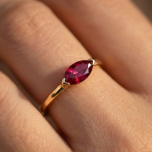 May include: A gold ring featuring a marquise-cut ruby gemstone. The ruby is a deep red color and is set in a gold band. The ring is shown on a finger.