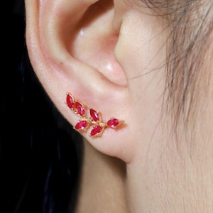 May include: Gold earrings with a branch design featuring red gemstones. The earrings are shown on a person's ear.