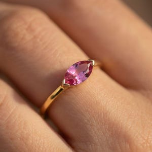 May include: A gold ring featuring a marquise-cut pink gemstone. The gemstone is set in a delicate gold band, showcasing a vibrant pink hue. The ring is a piece of jewelry.
