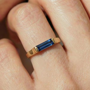 May include: A gold ring featuring a rectangular, deep blue gemstone. The ring's band is a simple, polished gold, with the gemstone set between two gold accents. The ring is shown on a person's finger.