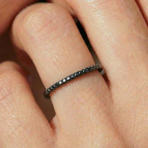 May include: A slender black ring, adorned with a line of small, black gemstones. The ring is displayed on a finger, highlighting the elegant design and dark hue of the jewellery.