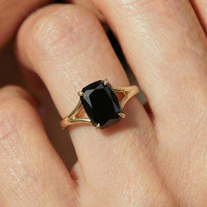 May include: A gold ring featuring a large, rectangular black gemstone. The ring's band has a split design, with the gemstone held in place by four prongs. The ring is shown on a person's finger.