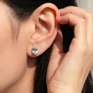 Puede incluir: Pendiente de botón con gema ovalada de color azul claro. El pendiente está engastado en un engaste de metal y se lleva en la oreja. La imagen muestra un primer plano de la oreja y el pendiente.