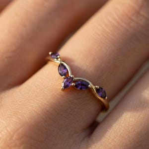 May include: A gold-colored ring featuring a curved design adorned with marquise-cut, deep purple gemstones. The ring's band curves gracefully, with the gemstones arranged in a leaf-like pattern. The ring is shown on a person's finger.