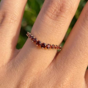 May include: A V-shaped ring crafted from gold metal, featuring a series of small, round, deep red garnet gemstones set along the band. The stones are arranged in a curved, chevron-like pattern that follows the contour of the metal band.